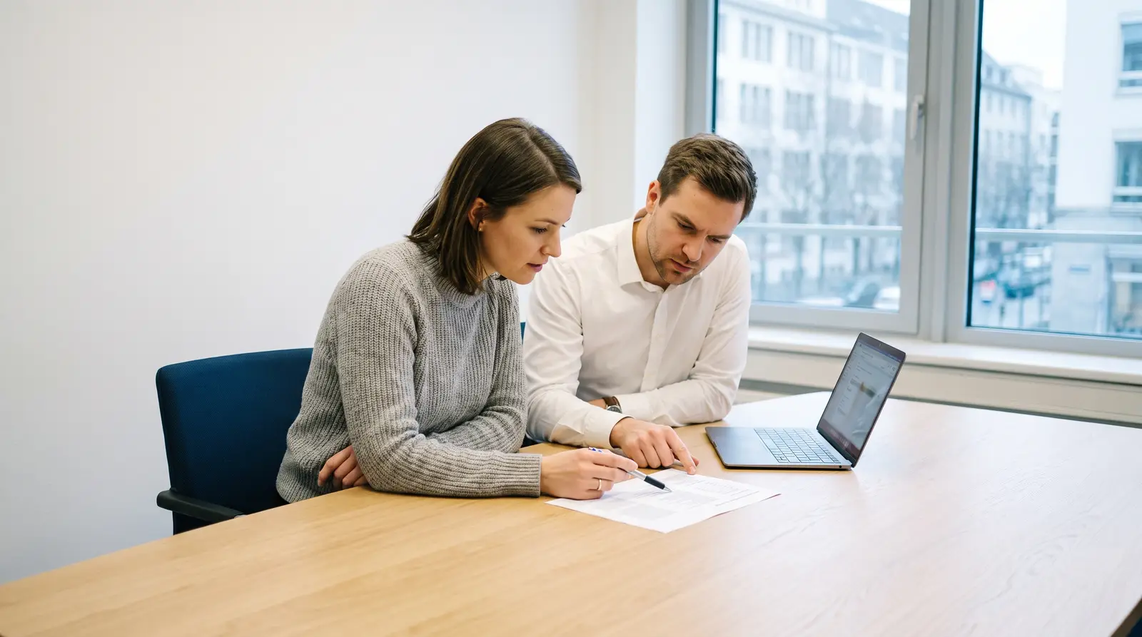 Two colleagues at a meeting table go through a printed sheet together, an open laptop beside them.